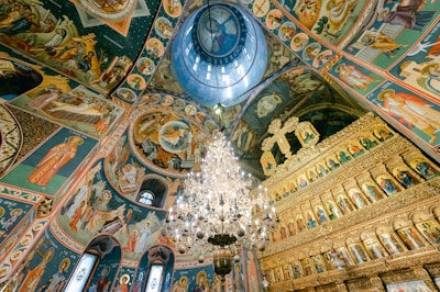 An opulent interior of a religious building featuring intricate frescoes covering the walls and ceiling. The artwork depicts various religious scenes and figures, while a large, ornate chandelier hangs prominently in the center. The architecture includes arches and domed structures with a color palette dominated by gold, blue, and earth tones.