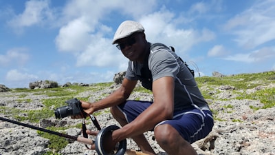A person wearing a gray hat and sunglasses is crouched on a rocky terrain with patches of green vegetation. They are dressed in a gray t-shirt and navy shorts, holding a DSLR camera in one hand and a fishing pole in the other, suggesting a leisurely activity. A backpack is slung over their shoulders. Above, the sky is bright with scattered white clouds.