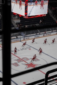 Hockey players wearing red uniforms are on an ice rink with a large electronic screen showing their images above. The rink is marked with a central logo and surrounded by advertising boards. Some spectators are visible, and the stands appear largely empty.