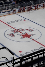 A group of ice hockey players in red uniforms is gathered near the side of an indoor hockey rink. The ice features a large logo with a star and Cyrillic letters at its center. The players appear to be preparing or practicing, with hockey sticks in hand. The surrounding area includes boards with advertisements and a few spectators in the stands.