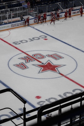 A group of ice hockey players in red uniforms is gathered near the side of an indoor hockey rink. The ice features a large logo with a star and Cyrillic letters at its center. The players appear to be preparing or practicing, with hockey sticks in hand. The surrounding area includes boards with advertisements and a few spectators in the stands.
