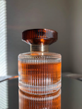 A glass perfume bottle with vertical ridges containing a warm amber liquid stands on a reflective surface. The cap is dark brown and has a ribbed texture, complementing the elegant design of the bottle.