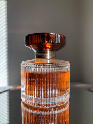 A glass perfume bottle with vertical ridges containing a warm amber liquid stands on a reflective surface. The cap is dark brown and has a ribbed texture, complementing the elegant design of the bottle.