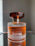 A glass perfume bottle with vertical ridges containing a warm amber liquid stands on a reflective surface. The cap is dark brown and has a ribbed texture, complementing the elegant design of the bottle.