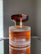 A glass perfume bottle with vertical ridges containing a warm amber liquid stands on a reflective surface. The cap is dark brown and has a ribbed texture, complementing the elegant design of the bottle.