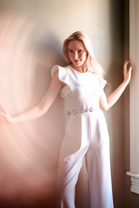 A person in a white outfit poses against a neutral-toned wall. The outfit is a stylish one-piece garment with decorative elements around the waist. Soft lighting creates a gentle, dreamy ambiance.