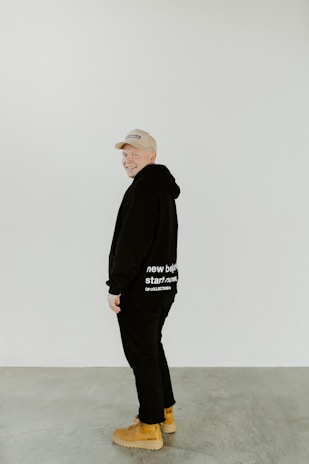 A person wearing a beige cap and black outfit stands in a minimalist setting. The black hoodie has white text that reads 'new beginning start now'. They are also wearing tan boots and smiling.