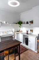 A cozy kitchen with wooden flooring features a dark wooden table and chair. The countertops are white, complementing the light-colored walls. There are open shelves with various plants, glass bottles, and dishes. A black trash bin is visible under the counter. A white sink is installed along the kitchen counter, and natural light brightens the room.