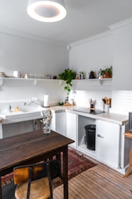 A cozy kitchen with wooden flooring features a dark wooden table and chair. The countertops are white, complementing the light-colored walls. There are open shelves with various plants, glass bottles, and dishes. A black trash bin is visible under the counter. A white sink is installed along the kitchen counter, and natural light brightens the room.