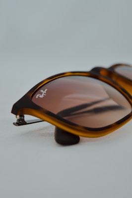 A close-up view of sunglasses with a tortoiseshell frame and brown-tinted lenses. The brand logo is visible on the top left corner of one lens. The background is a soft, light gray, emphasizing the product.