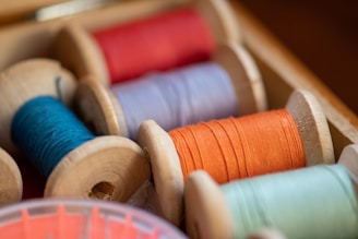A close-up view of several spools of thread in various colors, neatly arranged in a wooden box. The threads are wound tightly around wooden reels and display vibrant shades of red, orange, blue, and green. The box and spools have a slightly rustic appearance.