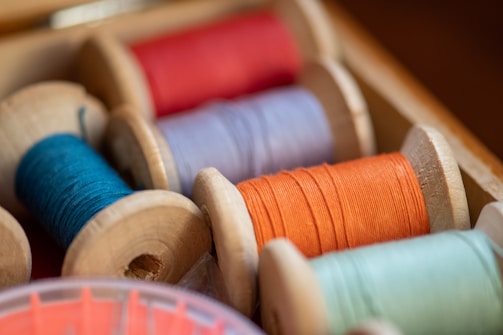 A close-up view of several spools of thread in various colors, neatly arranged in a wooden box. The threads are wound tightly around wooden reels and display vibrant shades of red, orange, blue, and green. The box and spools have a slightly rustic appearance.