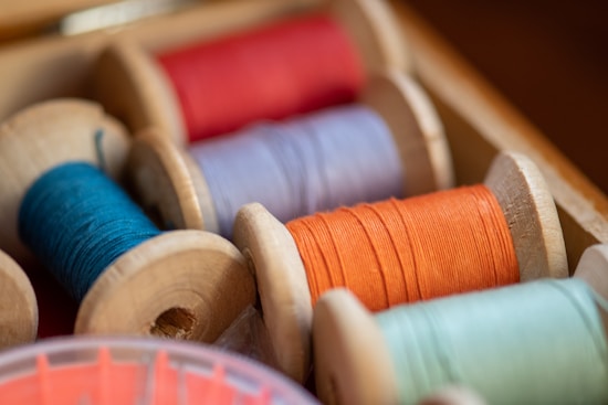 A close-up view of several spools of thread in various colors, neatly arranged in a wooden box. The threads are wound tightly around wooden reels and display vibrant shades of red, orange, blue, and green. The box and spools have a slightly rustic appearance.