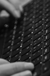 A close-up view of a person typing on a keyboard, with fingers pressing down on the keys. The image is in black and white, emphasizing the texture and details of the keyboard and the motion of typing.