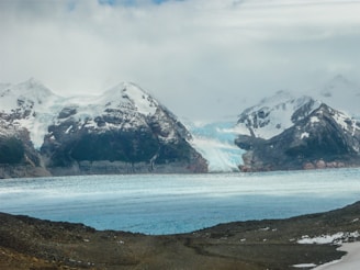 A vast glacier flows between rugged, snow-capped mountains under a cloudy sky, with a large expanse of ice at its base next to rocky terrain.