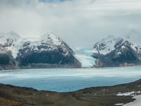 A vast glacier flows between rugged, snow-capped mountains under a cloudy sky, with a large expanse of ice at its base next to rocky terrain.