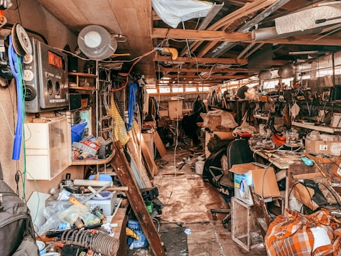 A cluttered workshop or garage filled with various tools, boxes, and equipment scattered across shelves and the floor. The space is crowded with old electronics, wooden boards, and hanging pieces of cloth. Overhead wiring and a mix of metal and wood construction give it a rustic and chaotic appearance.