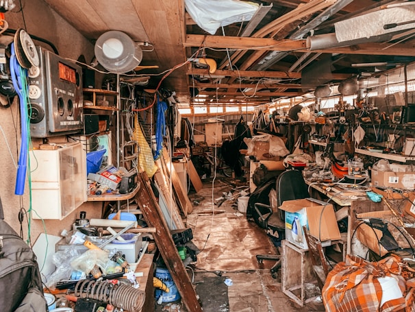 A cluttered workshop or garage filled with various tools, boxes, and equipment scattered across shelves and the floor. The space is crowded with old electronics, wooden boards, and hanging pieces of cloth. Overhead wiring and a mix of metal and wood construction give it a rustic and chaotic appearance.