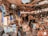 A cluttered workshop or garage filled with various tools, boxes, and equipment scattered across shelves and the floor. The space is crowded with old electronics, wooden boards, and hanging pieces of cloth. Overhead wiring and a mix of metal and wood construction give it a rustic and chaotic appearance.