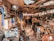 A cluttered workshop or garage filled with various tools, boxes, and equipment scattered across shelves and the floor. The space is crowded with old electronics, wooden boards, and hanging pieces of cloth. Overhead wiring and a mix of metal and wood construction give it a rustic and chaotic appearance.