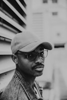 A black and white portrait features a man wearing glasses, a cap, and a denim jacket. The background shows an urban setting with blurred architecture.