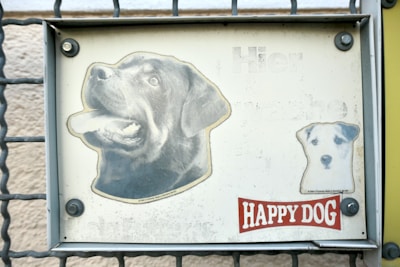 A metal sign attached to a gate features two printed dog images. The larger image depicts a black Labrador with its mouth open, and the smaller image shows a white dog with black spots. The background is off-white and appears slightly weathered. Below the images, there is a red and white label that reads 'HAPPY DOG'.