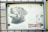 A metal sign attached to a gate features two printed dog images. The larger image depicts a black Labrador with its mouth open, and the smaller image shows a white dog with black spots. The background is off-white and appears slightly weathered. Below the images, there is a red and white label that reads 'HAPPY DOG'.