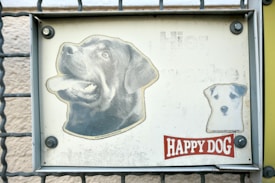 A metal sign attached to a gate features two printed dog images. The larger image depicts a black Labrador with its mouth open, and the smaller image shows a white dog with black spots. The background is off-white and appears slightly weathered. Below the images, there is a red and white label that reads 'HAPPY DOG'.