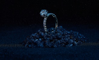 A diamond ring set on a small pile of dark, reflective stones against a black background. The ring is prominently adorned with diamonds, giving it a luxurious and elegant appearance. The lighting highlights the sparkle of the diamonds while the surrounding darkness enhances the ring's brilliance.