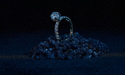 A diamond ring set on a small pile of dark, reflective stones against a black background. The ring is prominently adorned with diamonds, giving it a luxurious and elegant appearance. The lighting highlights the sparkle of the diamonds while the surrounding darkness enhances the ring's brilliance.