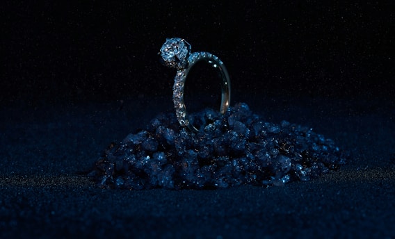 A diamond ring set on a small pile of dark, reflective stones against a black background. The ring is prominently adorned with diamonds, giving it a luxurious and elegant appearance. The lighting highlights the sparkle of the diamonds while the surrounding darkness enhances the ring's brilliance.