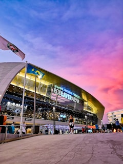 A modern sports arena with a large, illuminated sign bearing the name 'Rod Laver Arena.' A vibrant sky with purple and pink hues serves as the backdrop. A flag and several people can be seen outside the venue, adding a lively atmosphere.