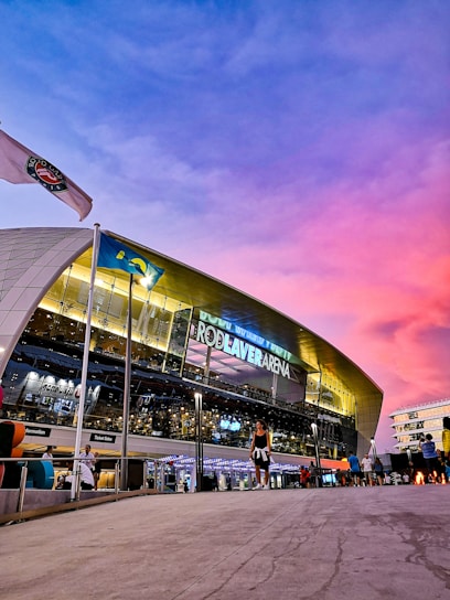 A modern sports arena with a large, illuminated sign bearing the name 'Rod Laver Arena.' A vibrant sky with purple and pink hues serves as the backdrop. A flag and several people can be seen outside the venue, adding a lively atmosphere.