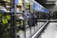 A long aisle with glass-door freezers in a supermarket, containing various frozen food items. The doors have handles, and there are informational signs above them. The area is well lit and spacious, creating a modern and clean atmosphere.