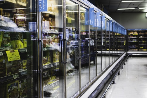 A long aisle with glass-door freezers in a supermarket, containing various frozen food items. The doors have handles, and there are informational signs above them. The area is well lit and spacious, creating a modern and clean atmosphere.