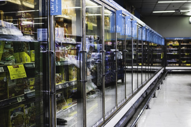 A long aisle with glass-door freezers in a supermarket, containing various frozen food items. The doors have handles, and there are informational signs above them. The area is well lit and spacious, creating a modern and clean atmosphere.