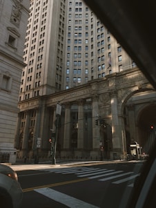 A grand, historic building with ornate architectural details, tall columns, and numerous windows stands majestically in an urban setting. The facade features classical designs and the words 'New Amsterdam MDCXXV'. The area seems quiet with a few street signs, a traffic light, and part of a crosswalk visible.
