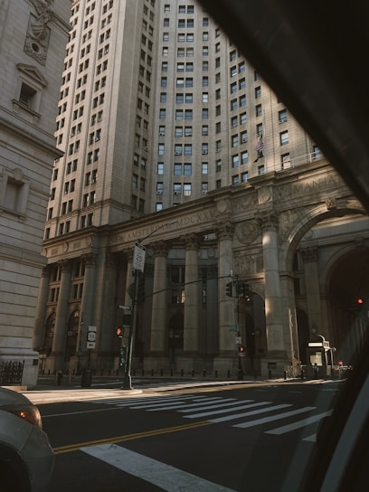 A grand, historic building with ornate architectural details, tall columns, and numerous windows stands majestically in an urban setting. The facade features classical designs and the words 'New Amsterdam MDCXXV'. The area seems quiet with a few street signs, a traffic light, and part of a crosswalk visible.
