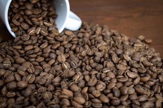A white mug is tipped over, spilling a large amount of roasted coffee beans onto a wooden surface. The beans are medium to dark brown, indicating a rich roast, and their glossy surfaces suggest freshness.