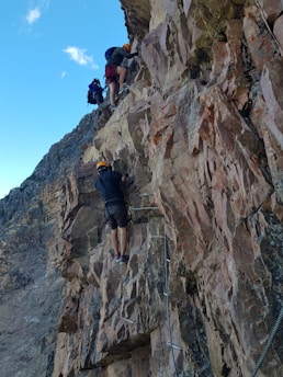People are climbing a steep, rocky mountain using metal steps and safety ropes. The cliff face appears rugged and challenging. Climbers are wearing helmets and backpacks, suggesting an organized climbing expedition. The sky is clear with a few clouds, indicating good weather.