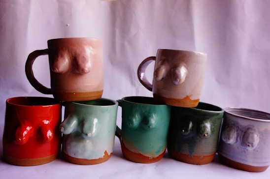 A collection of uniquely designed ceramic mugs, each featuring a raised, abstract sculpture on the side resembling human torsos. The mugs are arranged in two rows against a plain backdrop, showcasing various colors including pink, red, green, and grey.