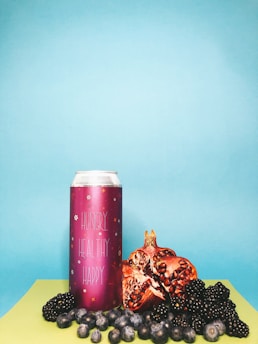 A vibrant composition with a tall pink can labeled 'Hungry Healthy Happy' surrounded by fresh fruit, including pomegranate halves, blackberries, and blueberries, set against a backdrop of light blue and lime green surfaces.