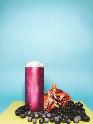 A vibrant composition with a tall pink can labeled 'Hungry Healthy Happy' surrounded by fresh fruit, including pomegranate halves, blackberries, and blueberries, set against a backdrop of light blue and lime green surfaces.
