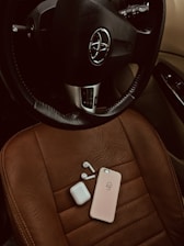 A black leather steering wheel with a Toyota logo is positioned above a brown leather car seat. On the seat, there is a pink phone case, a pair of wireless earbuds, and a white charging case arranged neatly.