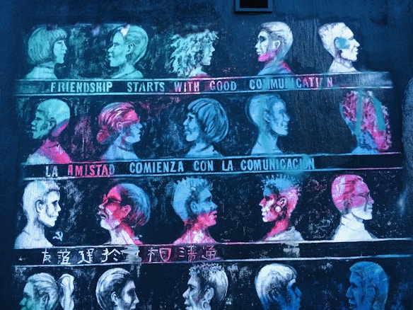 A mural features rows of stylized profile portraits in various vibrant colors. Each row is accompanied by a phrase about communication in different languages, including English, Spanish, and Chinese. The artwork uses contrast between bright neon colors and a dark background to highlight the faces.