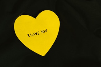A yellow heart-shaped note with the words 'I LOVE YOU' written in black is placed on a dark background.