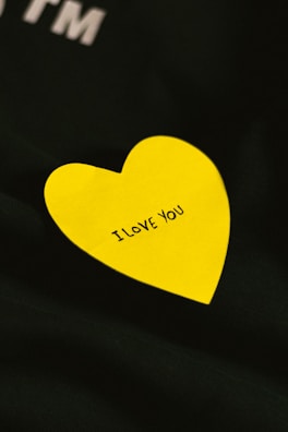A bright yellow heart-shaped note with the words 'I LOVE YOU' written in black, placed on a dark fabric background.