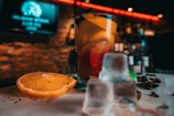 A close-up of a refreshing cocktail served in a glass with an orange slice garnish. Ice cubes are scattered on a wooden surface, adding to the chilled vibe. The background features a dimly lit bar with blurred shelves and a neon sign, contributing to a relaxed and inviting atmosphere.