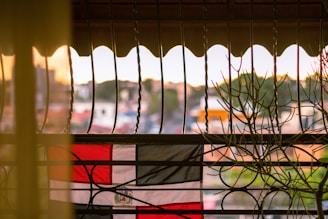 A flag with red, white, and black horizontal stripes is viewed through a decorative metal railing. The scene is backlit by a soft, warm sunset, with hints of green foliage visible in the blurred background.