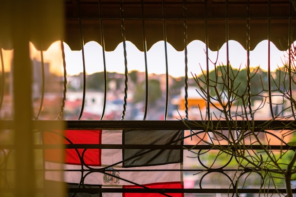 A flag with red, white, and black horizontal stripes is viewed through a decorative metal railing. The scene is backlit by a soft, warm sunset, with hints of green foliage visible in the blurred background.
